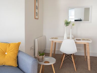 A dining area with a white table and chairs a grey sofa with a yellow cushion and a small round side table with a plant at The Towers - Llyn in Criccieth