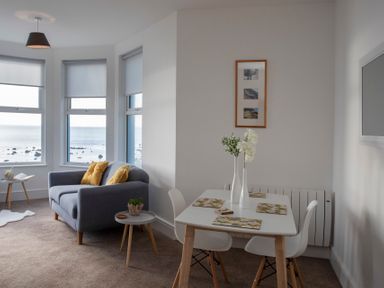A living area with a gray sofa and yellow cushions next to windows overlooking the sea and a white dining table with chairs and vases with flowers at The Towers - Llyn in Criccieth