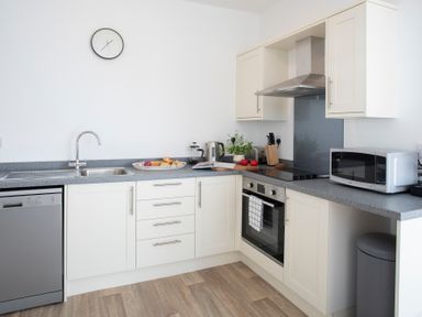 A kitchen with white cabinets gray countertops a stainless steel sink an oven a microwave and a clock on the wall at The Towers - Llyn in Criccieth