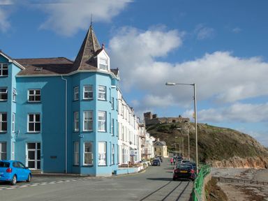 A street with a blue building and cars parked near a hill with castle ruins at The Towers - Llyn in Criccieth