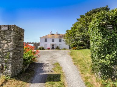 A driveway leading to a white two-story house with flowers and trees at Towyn Lodge in Trearddur Bay