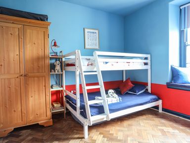 A bedroom with a white bunk bed blue and red walls a wooden wardrobe and a window seat at Towyn Lodge in Trearddur Bay