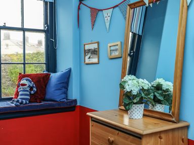 A corner with a window seat with cushions and a sock monkey toy beside a wooden dresser with a mirror and a potted plant at Towyn Lodge in Trearddur Bay