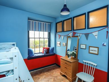 A blue and red children's bedroom with a bunk bed window seat and wooden dresser with mirror at Towyn Lodge in Trearddur Bay