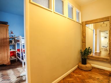 A hallway with a large wooden framed mirror and a plant on a wicker stool next to a doorway leading to a bedroom with bunk beds at Towyn Lodge in Trearddur Bay