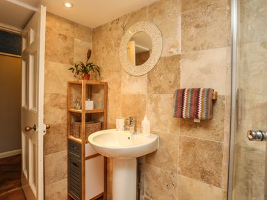A bathroom with a pedestal sink a round mirror towel rack with a striped towel wooden shelving unit with a plant and baskets at Towyn Lodge in Trearddur Bay