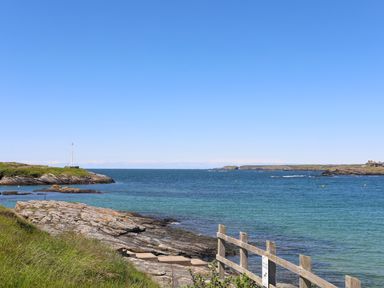 A coastal scene with rocky shoreline wooden railing and calm sea under a clear sky at Towyn Lodge in Trearddur Bay