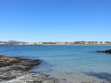 A coastal scene with rocky shore in the foreground and a town with houses across the water at Towyn Lodge in Trearddur Bay