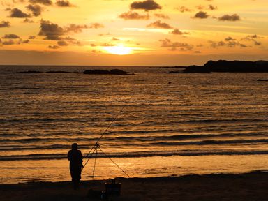 A person fishing with rods on a beach at sunset with rocks in the sea at Towyn Lodge in Trearddur Bay