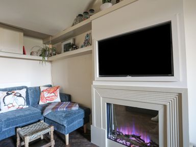 A corner of a living room with a blue sectional sofa with two decorative pillows a small woven stool a built in electric fireplace and a mounted flat screen TV with shelves with plants and framed pictures at Towyn Lodge in Trearddur Bay