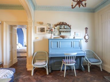 A room with a blue piano, two wicker chairs with cushions, a white chair, and framed pictures on the wall at Towyn Lodge in Trearddur Bay