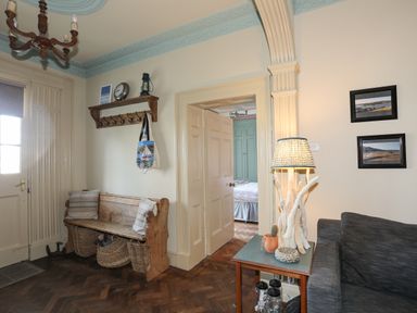 A seating area with a wooden bench and cushions under a coat rack next to a side table with a lamp and a dark sofa at Towyn Lodge in Trearddur Bay