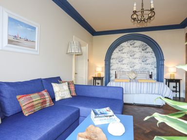 A room with a blue couch with pillows a bed with striped bedding under a blue arch and bedside tables with lamps at Towyn Lodge in Trearddur Bay