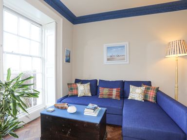A living room with a blue sofa and cushions a floor lamp a coffee table with books and a plant by the window at Towyn Lodge in Trearddur Bay