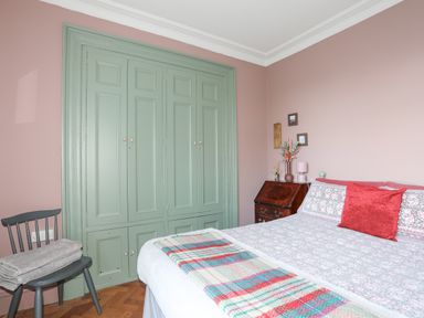 A bedroom with a bed a red cushion a colorful blanket a wooden chair with towels and green built-in wardrobes at Towyn Lodge in Trearddur Bay