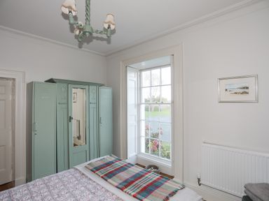 A bedroom with a bed covered with a patterned blanket and colorful throw a green wardrobe with a mirror large window with a view outside a radiator and a framed picture on the wall at Towyn Lodge in Trearddur Bay