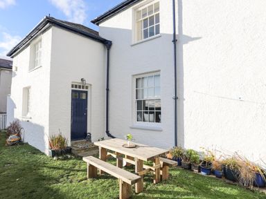 A white house exterior with a blue door and windows next to a wooden picnic table with benches and potted plants on the grass at Towyn Lodge in Trearddur Bay