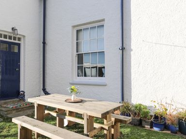 A wooden outdoor table with benches on grass in front of a white building with a blue door and a window with pillows in Towyn Lodge in Trearddur Bay