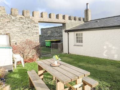 A garden with a wooden picnic table and benches a white building stone walls and a gate at Towyn Lodge in Trearddur Bay