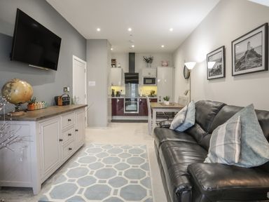 A living room with a black leather sofa and blue patterned rug next to a dining table and open kitchen at The Pot Loft in Benllech