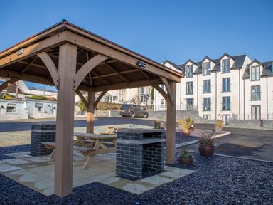 A wooden gazebo with picnic tables and a brick barbecue in a paved area outside a multi-story white building at The Pot Loft in Benllech