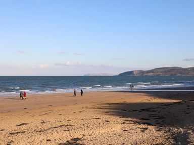 A sandy beach with five people walking near the water and a hillside in the background at The Pot Loft in Benllech