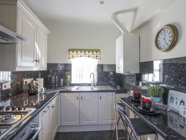 A kitchen with white cabinets black countertops a window with a polka dot valance and a small table with two cups and a plant at Tyn Towyn - Bwthyn Carreg in Trearddur Bay