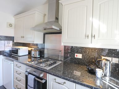 A kitchen with white cabinets black countertops a gas stove microwave toaster kettle and a cup on the counter at Tyn Towyn - Bwthyn Carreg in Trearddur Bay