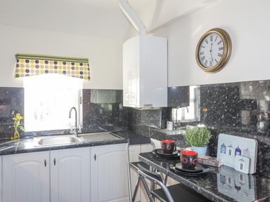 A kitchen with a sink under a window with polka dot curtains and a counter with two cups plates and a plant at Tyn Towyn - Bwthyn Carreg in Trearddur Bay