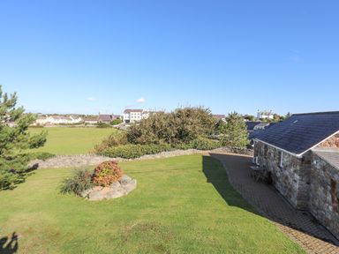 A grassy yard with a stone flower bed and a stone building with a paved walkway and houses in the background at Tyn Towyn - Bwthyn Carreg in Trearddur Bay
