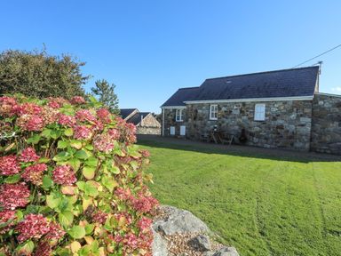A stone cottage with a slate roof a garden with green grass and pink flowers at Tyn Towyn - Bwthyn Carreg in Trearddur Bay