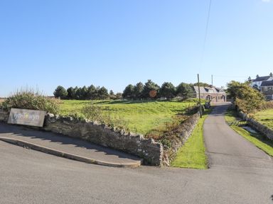A road leading to stone houses with a grassy field and trees at Tyn Towyn - Bwthyn Carreg in Trearddur Bay