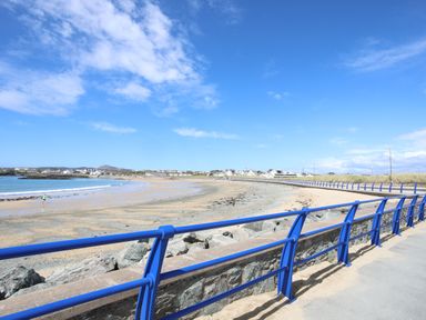 A beach with sand and rocks next to a walkway with blue railings and houses in the distance at Tyn Towyn - Bwthyn Carreg in Trearddur Bay