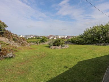 A grassy backyard with shrubs rocks and a stone wall with houses in the background in Trearddur Bay