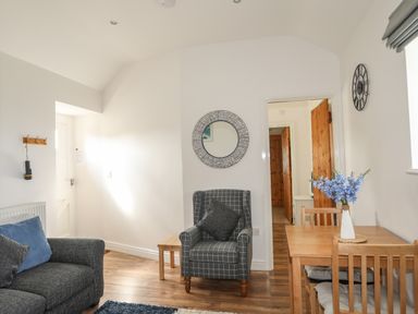 A living room with a gray armchair a wooden table with chairs and flowers and a sofa at Tyn Towyn - Bwthyn Carreg in Trearddur Bay