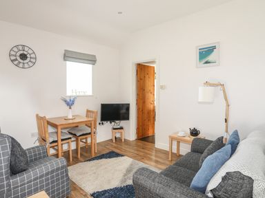 A living room with a gray sofa and armchair a small dining table with two chairs a wall-mounted tv and wooden furniture at Tyn Towyn - Bwthyn Carreg in Trearddur Bay