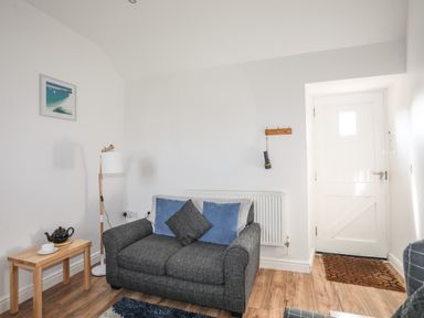 A sitting area with a gray sofa with cushions a wooden side table with a teapot and cup a floor lamp a white door and wooden flooring at Tyn Towyn - Bwthyn Carreg in Trearddur Bay