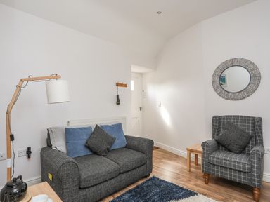 A living room with a grey sofa and armchair a wooden floor and a round mirror on the wall at Tyn Towyn - Bwthyn Carreg in Trearddur Bay