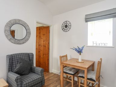 A room with a wooden dining table and two chairs a grey checkered armchair a round wall mirror a clock and a window with a Home sign at Tyn Towyn - Bwthyn Carreg in Trearddur Bay