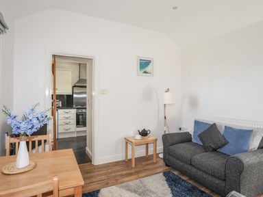 A living room with a gray sofa cushions a small wooden table with a teapot a floor lamp a wooden dining table with blue flowers and a view into a kitchen at Tyn Towyn - Bwthyn Carreg in Trearddur Bay