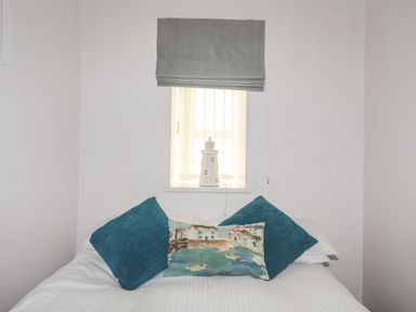 A bed with three pillows under a small window with a gray blind and a lighthouse decoration on the window sill at Tyn Towyn - Bwthyn Carreg in Trearddur Bay