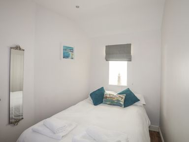 A bedroom with a bed with white linens and pillows and a small window with a blind and a mirror on the wall at Tyn Towyn - Bwthyn Carreg in Trearddur Bay