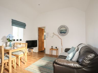 A living room with a brown leather sofa, dining table with chairs, and a television at Tyn Towyn - Bwthyn Haf in Trearddur Bay