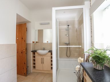 A bathroom with a wooden door, a sink with cabinet, a shower area, and a window with plants on the counter at Tyn Towyn - Bwthyn Haf in Trearddur Bay