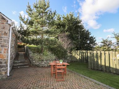 An outdoor patio with a wooden table and chairs next to a stone building and garden at Tyn Towyn - Bwthyn Haf in Trearddur Bay