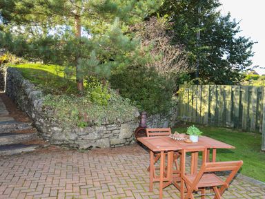 An outdoor patio area with a wooden table and chairs next to a stone wall and green plants at Tyn Towyn - Bwthyn Haf in Trearddur Bay