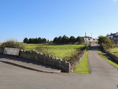 A driveway leading to stone buildings with a grass field and trees at Tyn Towyn - Bwthyn Haf in Trearddur Bay