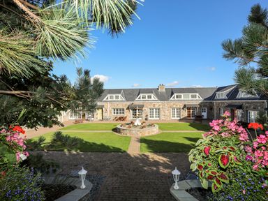 A courtyard with a central fountain and lawn surrounded by stone buildings with dormer windows at Tyn Towyn - Ty Glas in Trearddur Bay