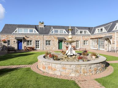 A courtyard with a stone fountain surrounded by grass and a stone building with multiple windows and doors at Tyn Towyn - Ty Glas in Trearddur Bay