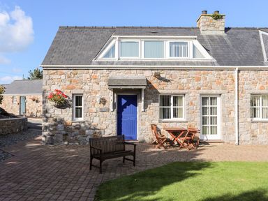 Stone house with blue door wooden bench and table with chairs on paved patio at Tyn Towyn - Ty Glas in Trearddur Bay